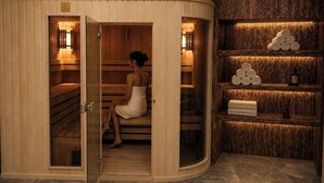 A sauna with wooden benches, a woman sitting, and shelves with towels and bottles. - Eighty Eight Hotel and Spa (Tsaghkadzor)