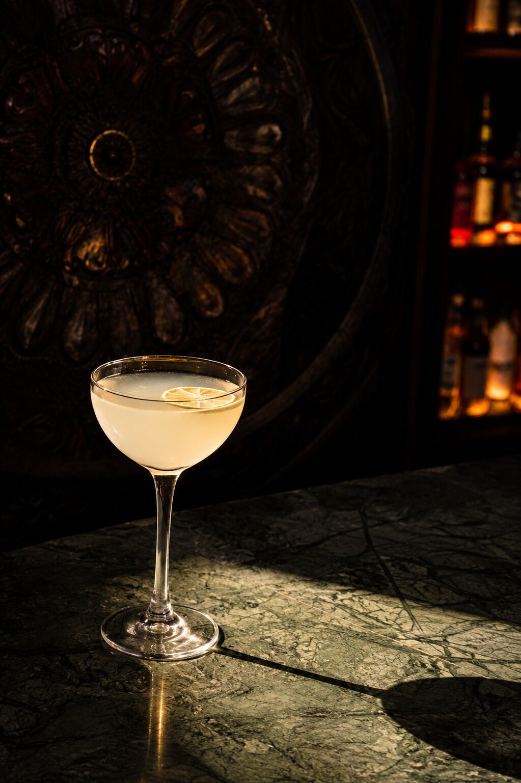 A cocktail in a glass on a marble surface with a decorative background and bottles in the background.