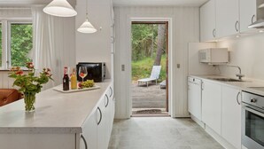 Une cuisine moderne avec des meubles blancs, un grand îlot central agrémenté d’une bouteille de vin et de verres, et une vue sur une terrasse en bois et la verdure environnante.