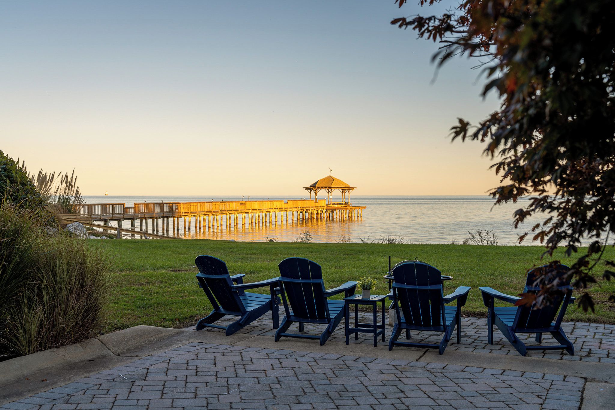 A waterfront view with a pier and a gazebo. There are four Adirondack chairs and a table on the patio.