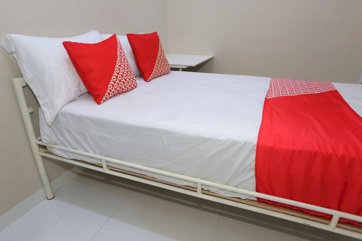 Ein Zimmer mit einem Einzelbett, weißer Bettwäsche, zwei roten Kissen mit weißem Muster und einer roten Decke.
