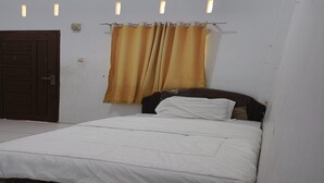 Un lit simple avec un matelas blanc et un couvre-lit à motifs, une porte marron et un rideau d’une teinte jaunâtre.