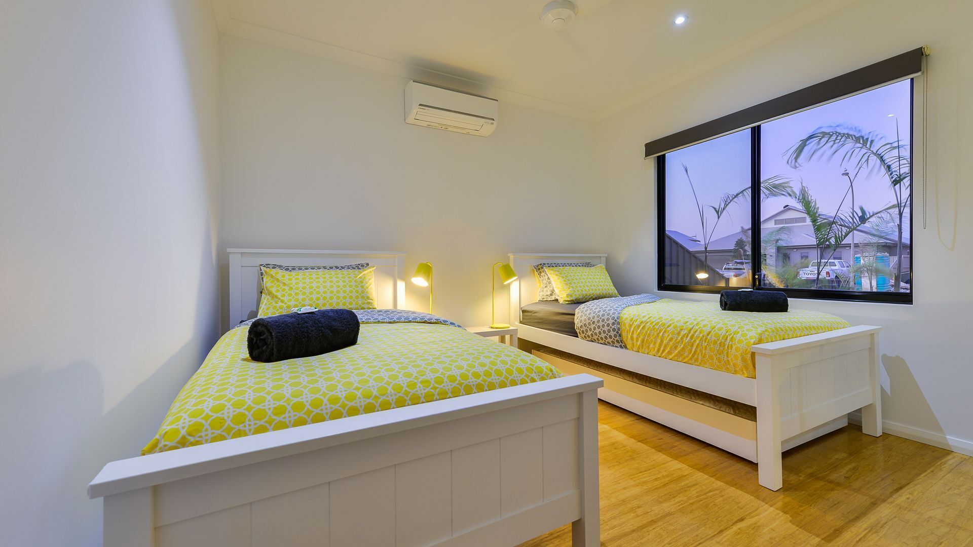 A bedroom with two beds, each with a patterned bedspread and matching pillows, a window with a view of palm trees, and an air conditioning unit on the wall.