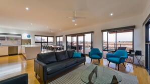 Una sala moderna con un sofá de cuero negro, una mesa de centro de vidrio y dos sillones azules. Cuenta con una zona de cocina con lavavajillas y un ventanal con vistas a una terraza.