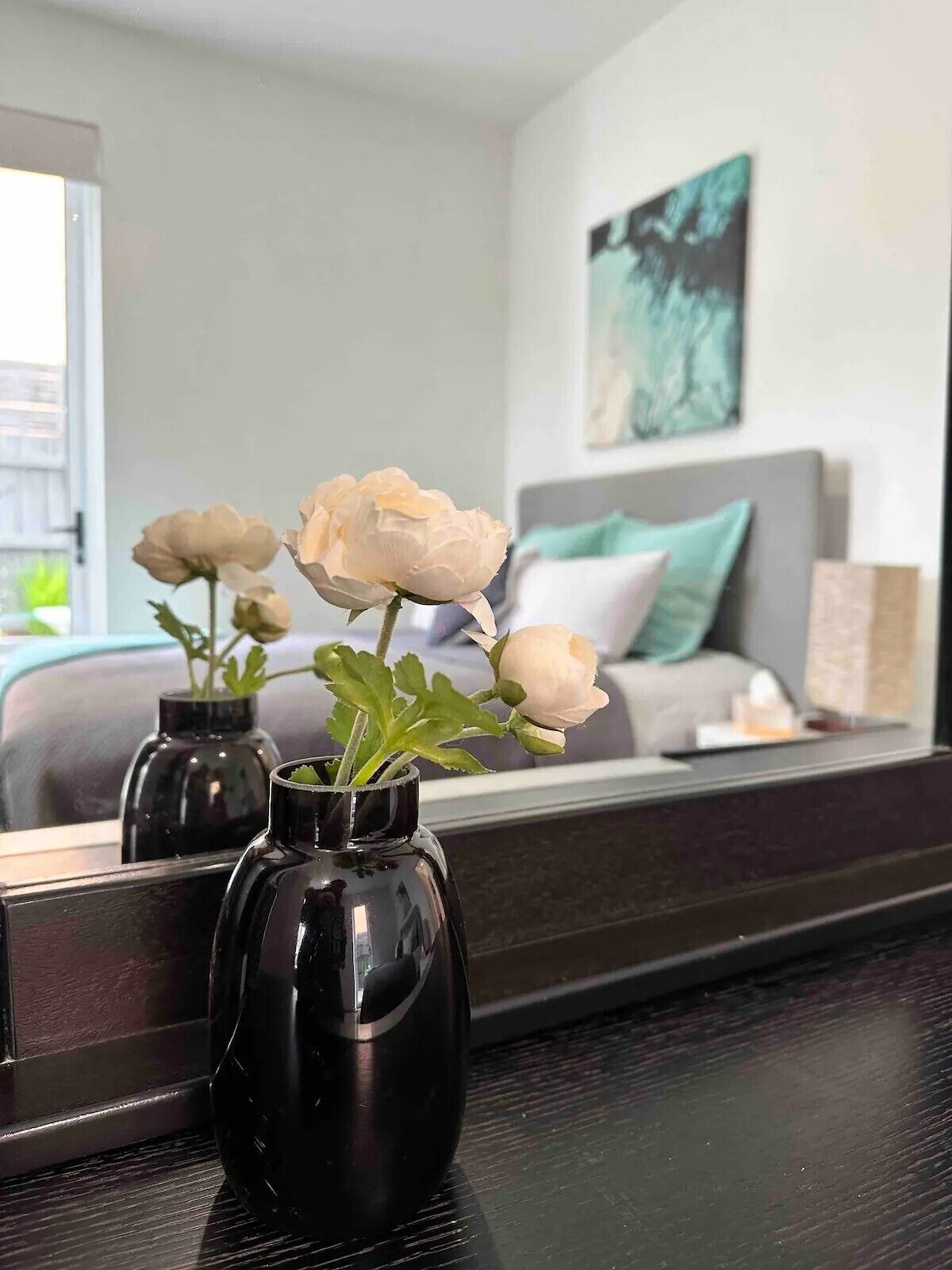 Quarto de hotel com um espelho grande refletindo a cama e um quadro na parede. Há dois vasos pretos com flores brancas sobre uma mesa em primeiro plano.