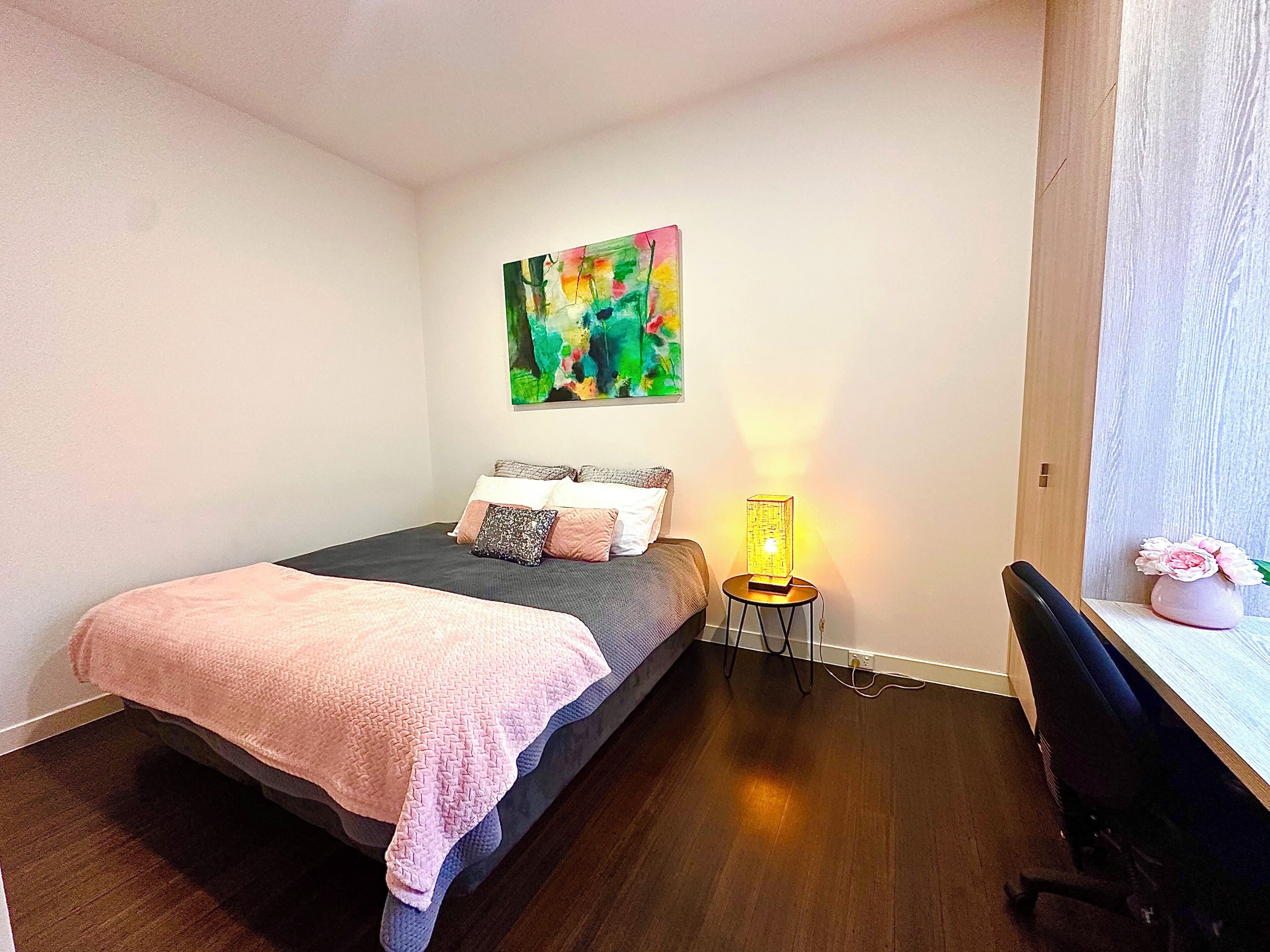 A bedroom with a bed, a painting on the wall, a bedside lamp, and a desk with a vase.