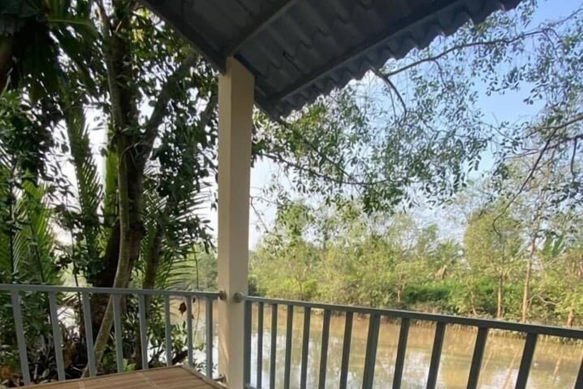 A covered outdoor area with a bamboo table and two chairs, a tiled floor, and a railing overlooking a body of water.