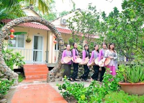 A group of people standing in a garden with a stone archway, a house with a red roof, and a staircase leading to the entrance. - Nghenh Xuan Farm - WellSpring Farm  (Chau Thanh)