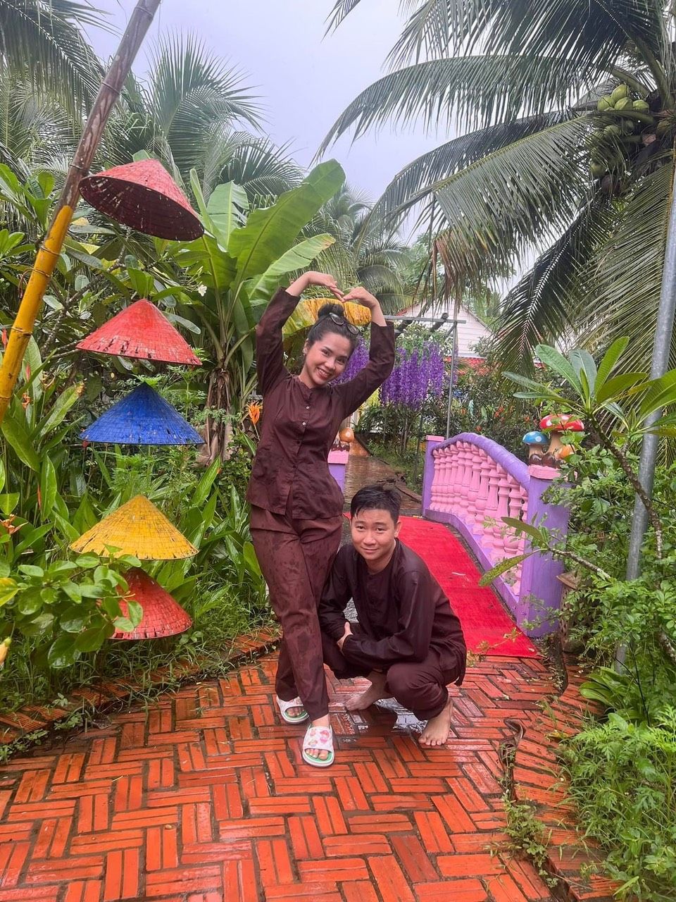 A person standing with a red umbrella, another person crouching, and colorful umbrellas in a garden with a brick pathway.