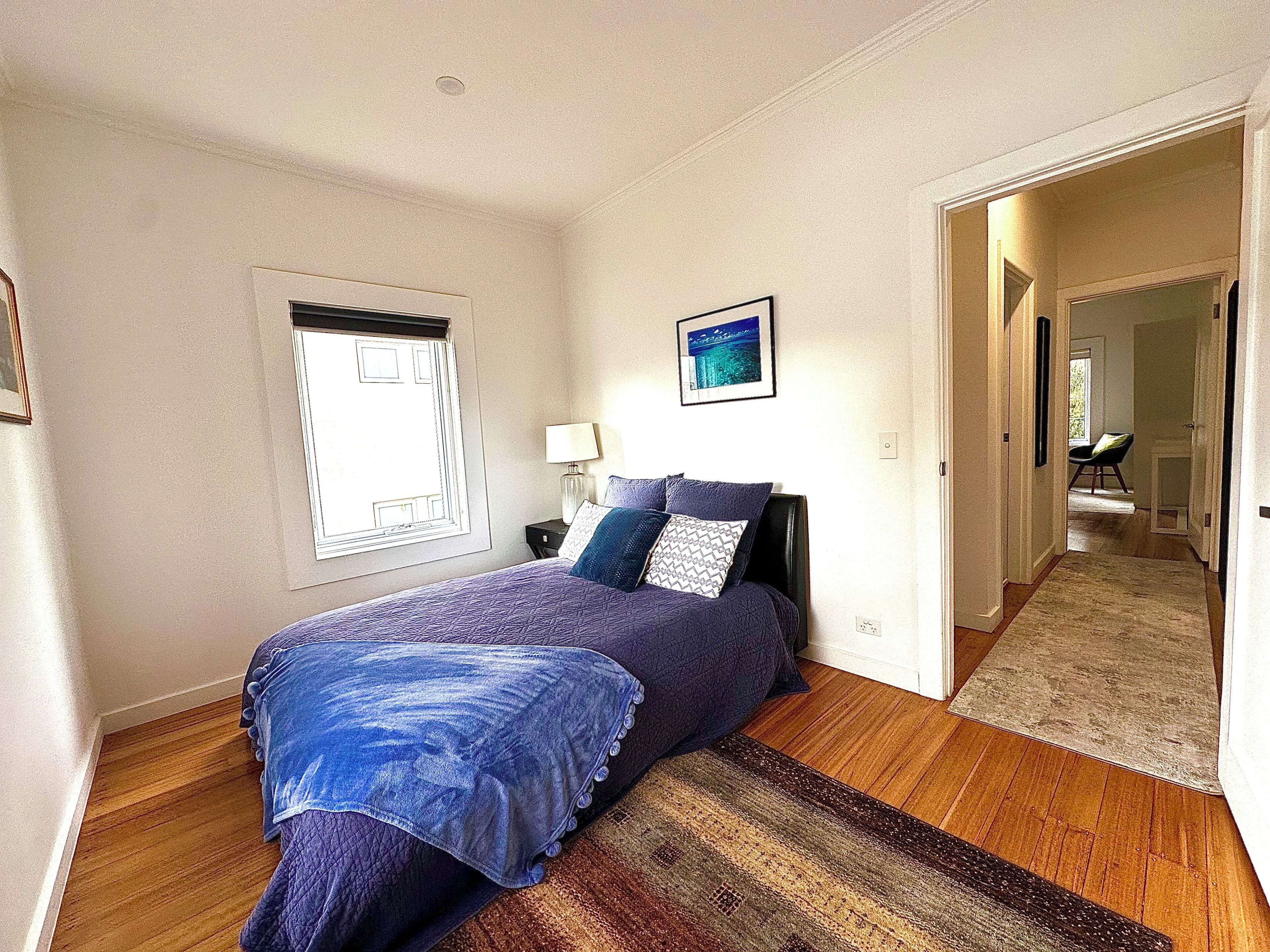 A bedroom with a bed, a nightstand, a window, a door, and a hallway leading to another room.