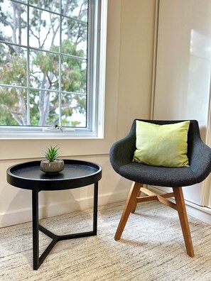 Una habitación con ventana, una pequeña mesa redonda con una planta y una silla moderna con cojín.