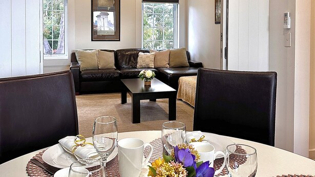 A dining area with a table set for four, a coffee table with a vase of flowers, and a view into a living room with a sofa and a window. - Luxury & Grandeur | Balcony Views| Prime Location, Parking (Hawthorn)