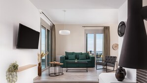A modern living room with a green sofa, a coffee table, a TV mounted on the wall, and a staircase leading to a balcony. - Cavos Boutique Homes (Corinth)