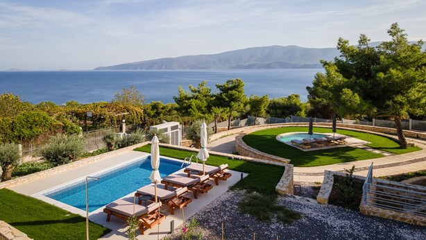 A swimming pool with lounge chairs and umbrellas, surrounded by a well-maintained lawn and trees, with a view of the sea and mountains in the background. - Cavos Boutique Homes (Corinth)