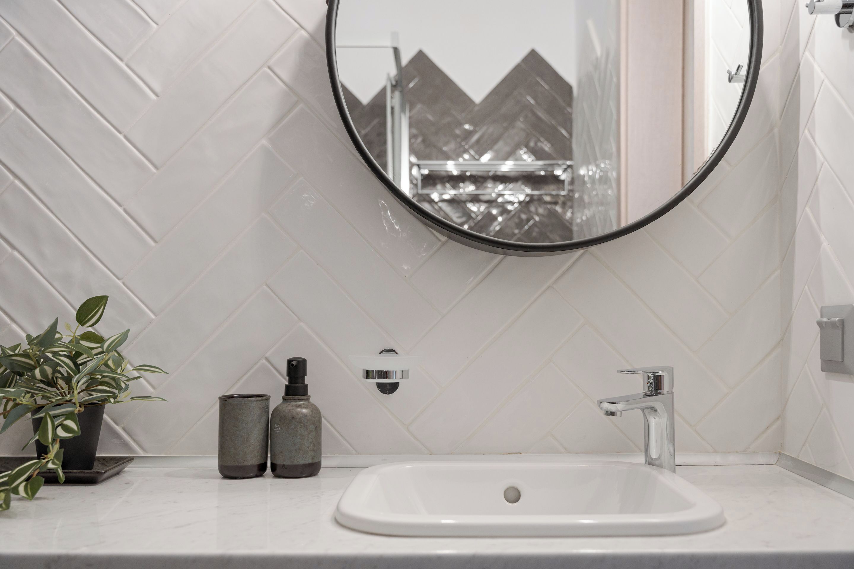 A bathroom with a white herringbone tile backsplash, a round mirror, a white sink, a modern faucet, a potted plant, and two bottles on the countertop.