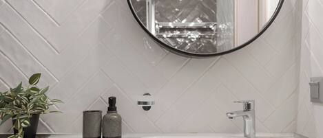 A bathroom with a white herringbone tile backsplash, a round mirror, a white sink, a modern faucet, a potted plant, and two bottles on the countertop.
