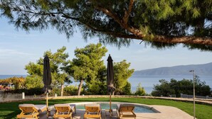 A poolside area with lounge chairs and umbrellas, surrounded by trees and a view of the sea. - Cavos Boutique Homes (Corinth)