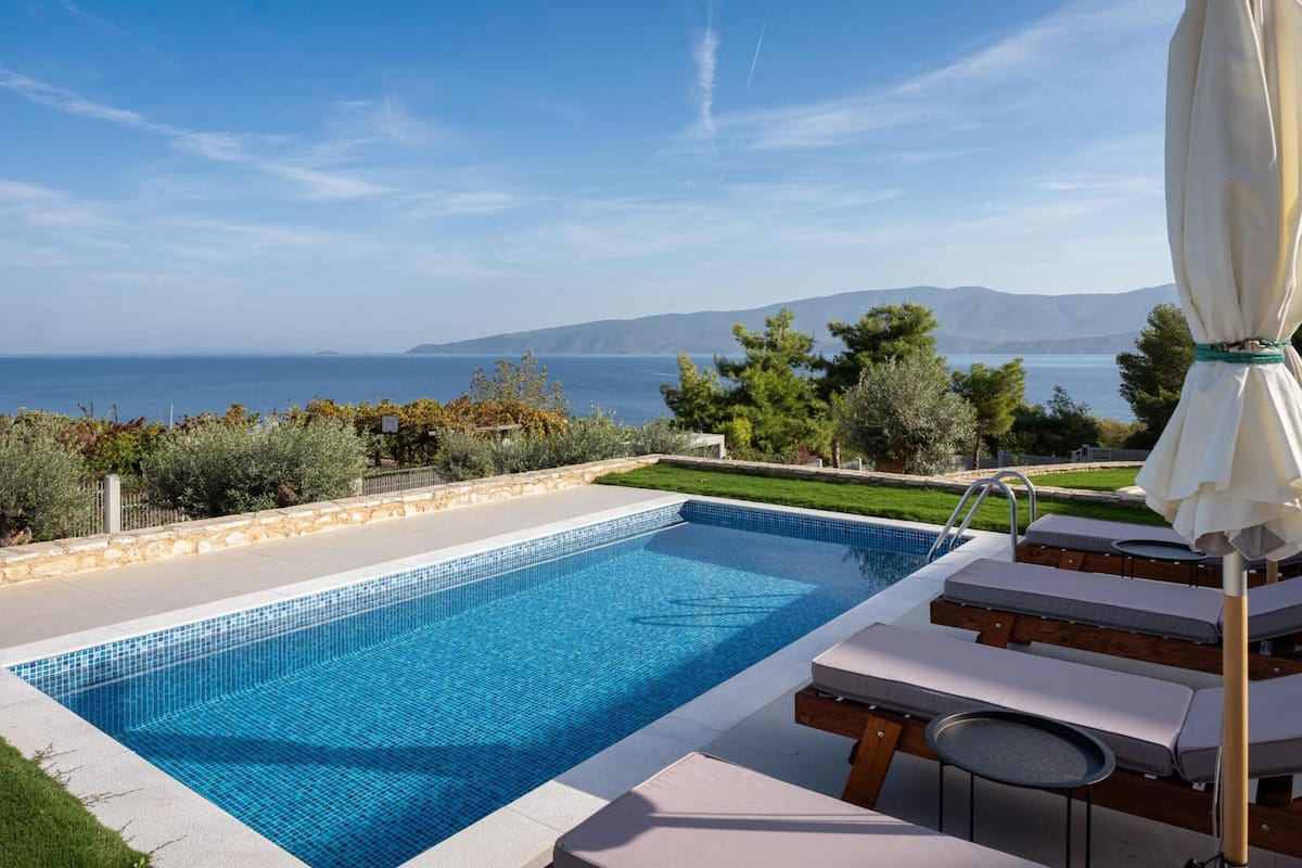 A rectangular swimming pool with clear blue tiles, surrounded by lounge chairs and a small table.