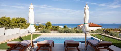 A poolside area with lounge chairs and umbrellas, a clear view of the sea, and a well-maintained lawn.