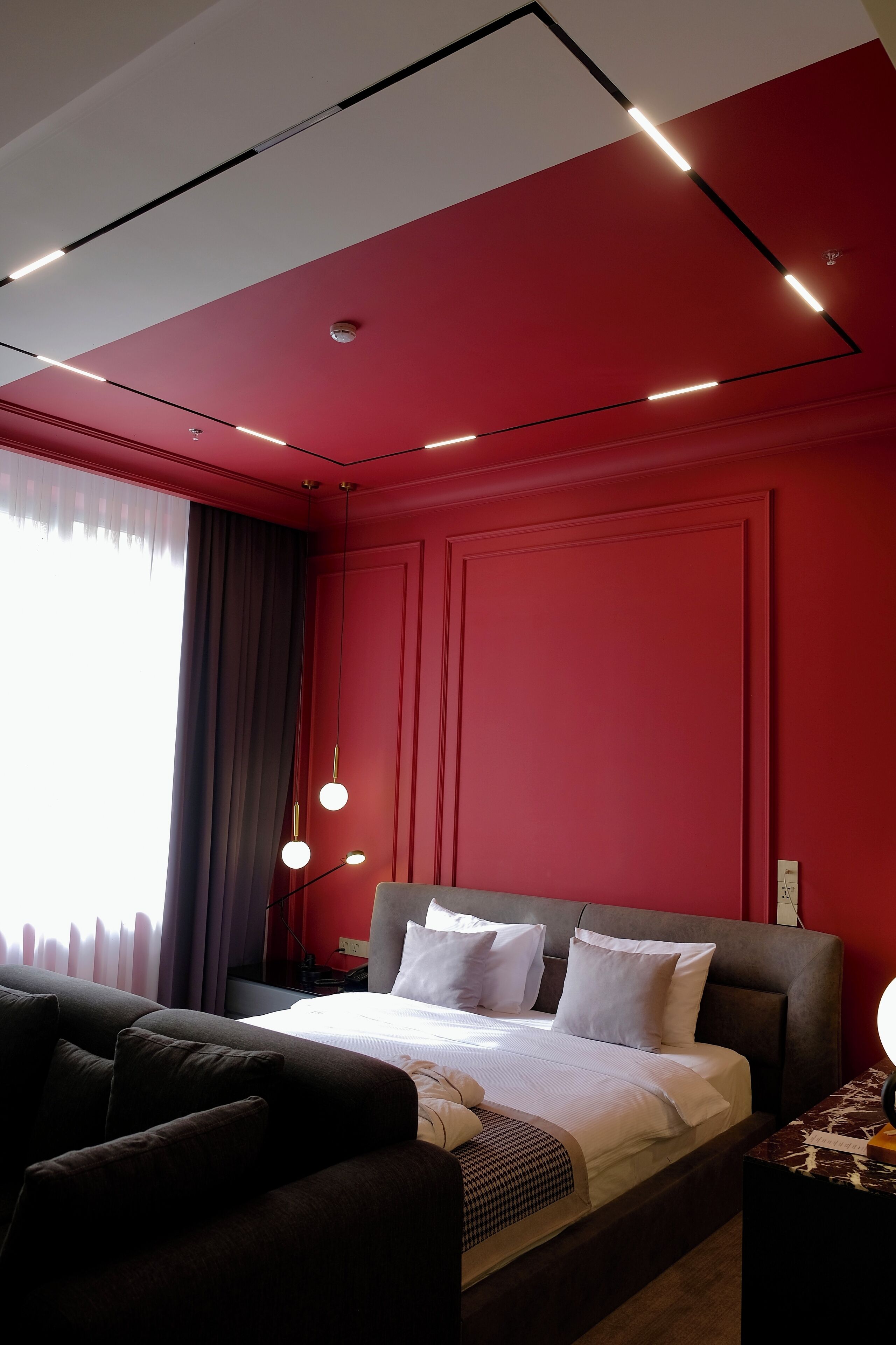 Ein modernes Hotelzimmer mit roter Akzentwand, einem großen Bett mit weißer Bettwäsche, einer dunklen Couch und einem Nachttisch mit Lampe.