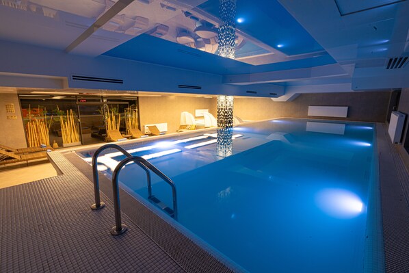 An indoor swimming pool with a clear water surface, a metal ladder, and a ceiling with recessed lighting.