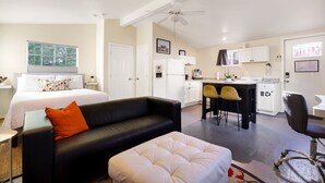A modern hotel room with a bed, a black leather sofa, a white ottoman, a dining area with a kitchen, and a ceiling fan.