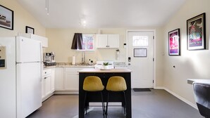 A compact kitchen with white cabinets, a refrigerator, a sink, and a window with a curtain. There are two yellow chairs at a small table.