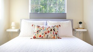 A neatly made bed with a white quilted bedspread and two pillows, one with a colorful floral pattern. There is a window above the bed with a view of greenery outside. Two bedside tables with lamps are visible on either side of the bed.