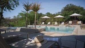 Un área junto a la piscina con sillones de descanso, sombrillas y un sombrero en un banco.