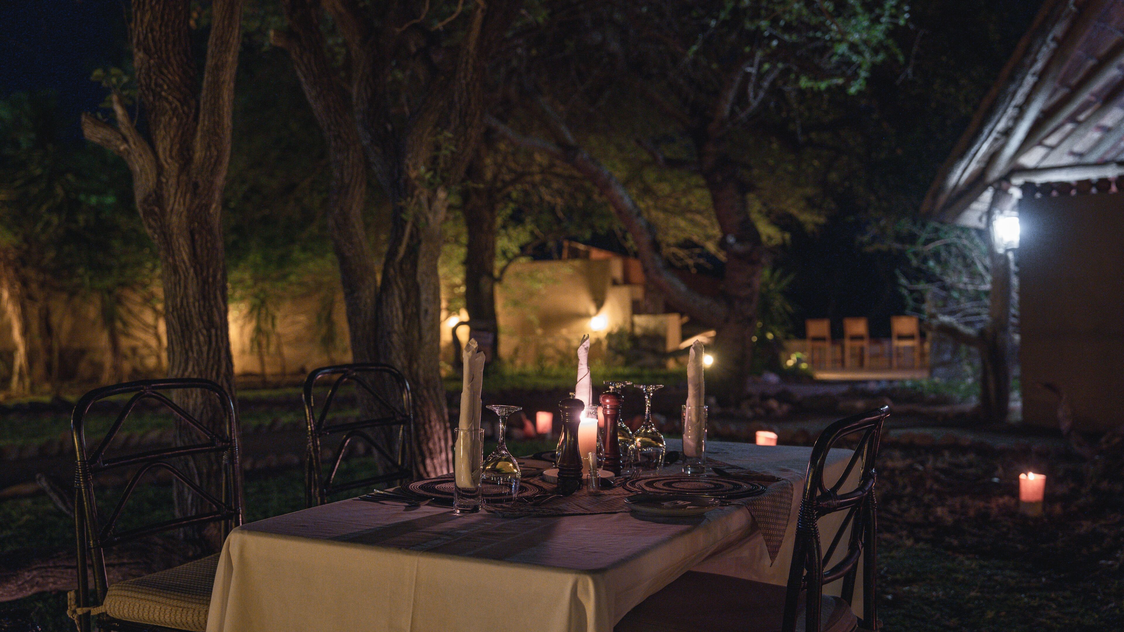 Un espace de restauration en plein air avec une table dressée pour deux personnes, entourée d’arbres et avec un bâtiment en arrière-plan.