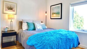 Une chambre à coucher avec un lit, une table de chevet avec une lampe, un tableau encadré au mur et une fenêtre donnant sur un paysage.