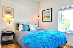 A bedroom with a bed, a nightstand with a lamp, a framed picture on the wall, and a window with a view.