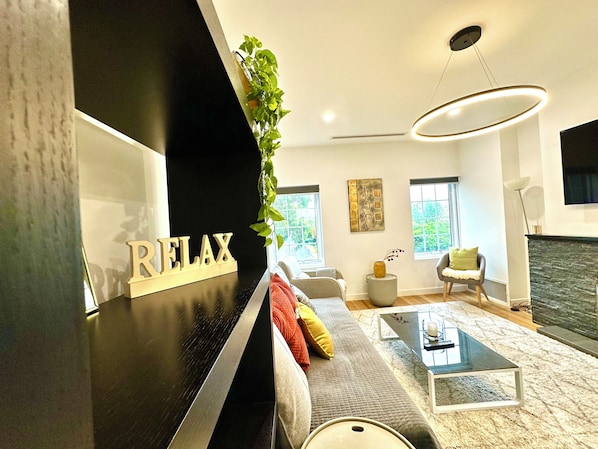 A modern living room with a black piano, a sofa with colorful cushions, a glass coffee table, a fireplace, and a large window.