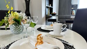 Une table à manger est dressée avec des assiettes blanches, une serviette pliée, un verre d’eau et un verre de vin. Un vase rempli de fleurs et un saladier de fruits sont posés sur la table.