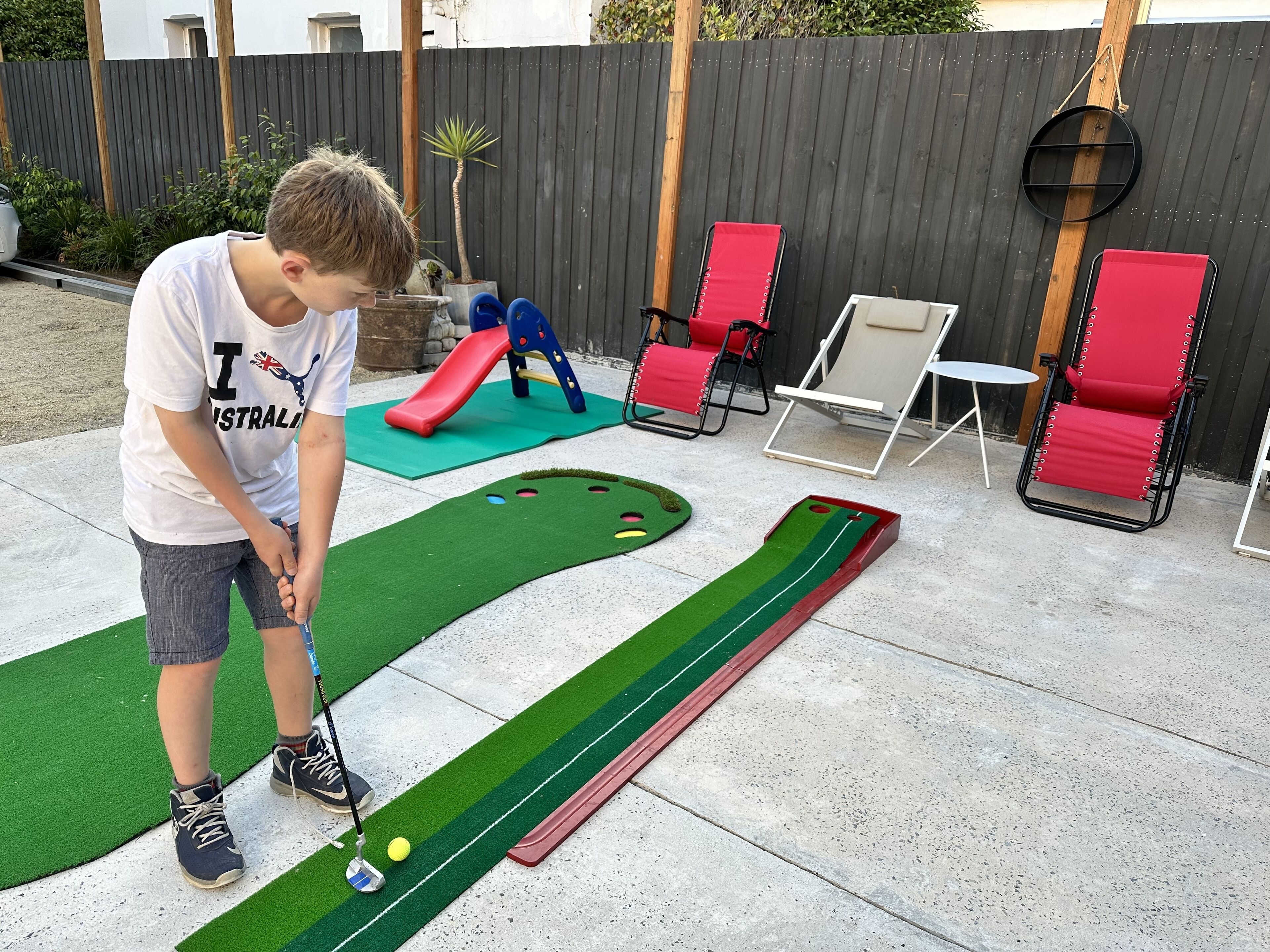 A child playing mini-golf in a backyard with a slide, red chairs, and a wooden fence.