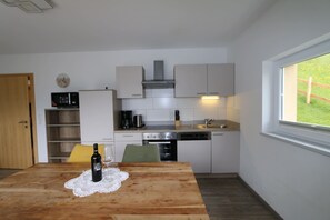 Una cocina compacta con armarios blancos, un microondas, una estufa y un lavavajillas. Hay una mesa de madera con una botella y una copa, y una ventana con vistas a la vegetación.