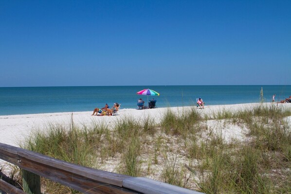 A beach with people relaxing under umbrellas, a wooden fence, and sand dunes.