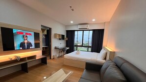 A modern hotel room with a large TV displaying 'The Idol', a bed, a sofa, a desk, and a view of the city through large windows. - JIA HAUS Hotel Apartment (Buriram)
