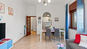 A modern living room with a dining area, a kitchen in the background, and a large window with curtains.