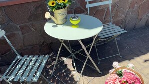 An outdoor patio with a round table, two metal chairs, a potted plant with yellow flowers, and a basket of pink flowers.