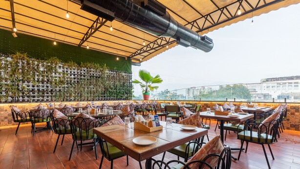 An outdoor restaurant with wooden tables and chairs, a green wall with plants, and a city view in the background. - HOTEL KALINGA (Indore)