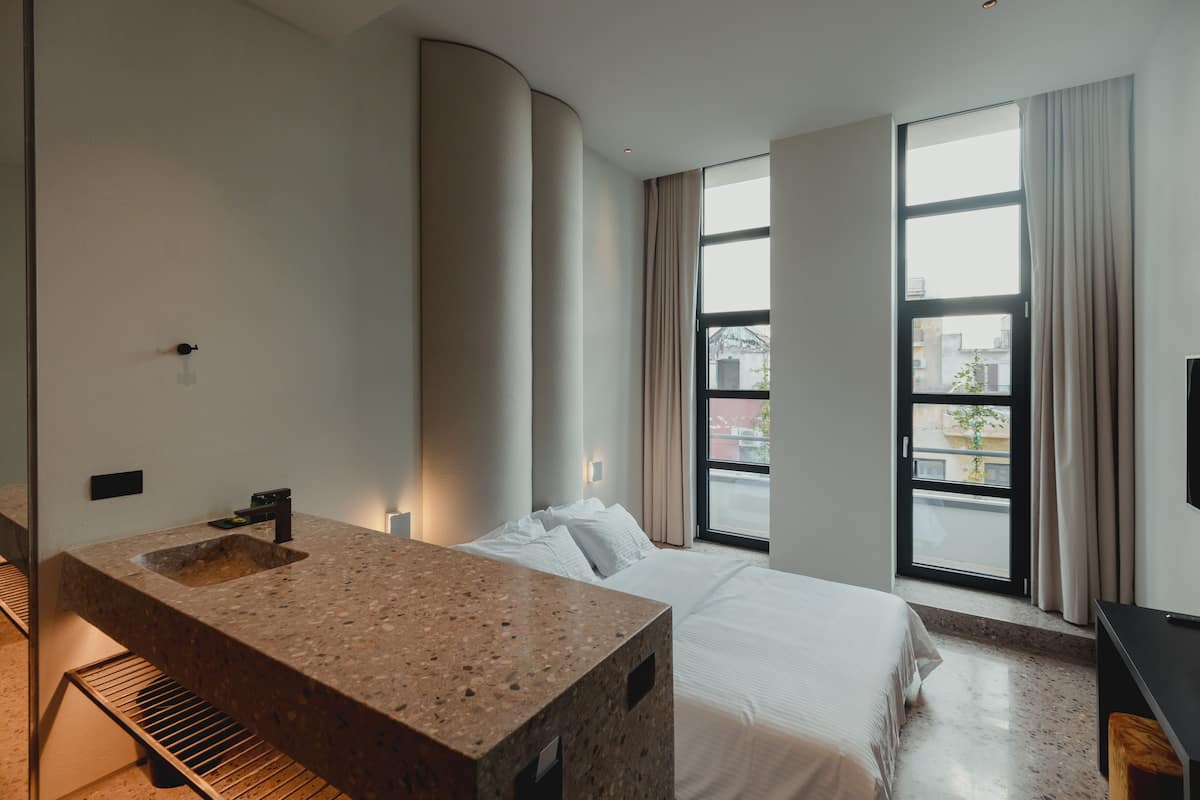 A modern hotel room with a large bed, a built-in granite sink, and large windows offering a city view.