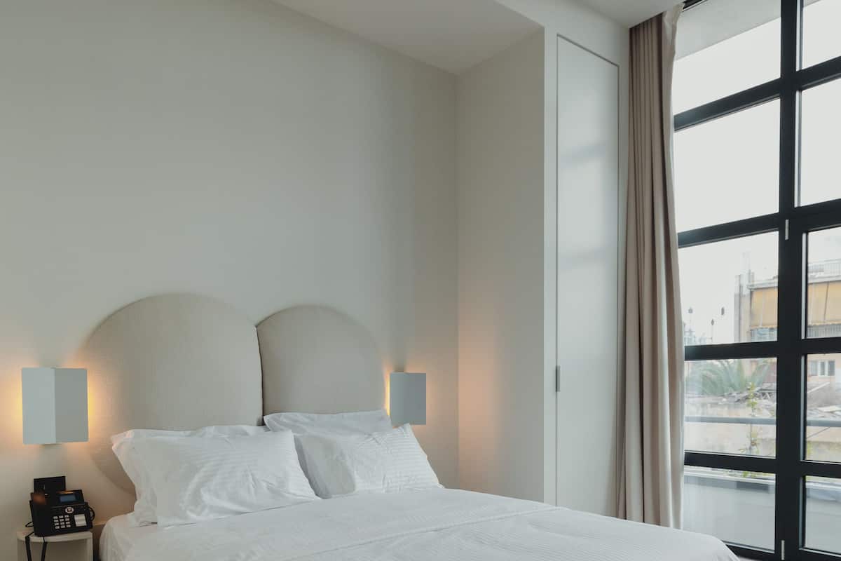 A modern hotel room with a large bed, two bedside lamps, a phone on the nightstand, and a large window with a view of the city.