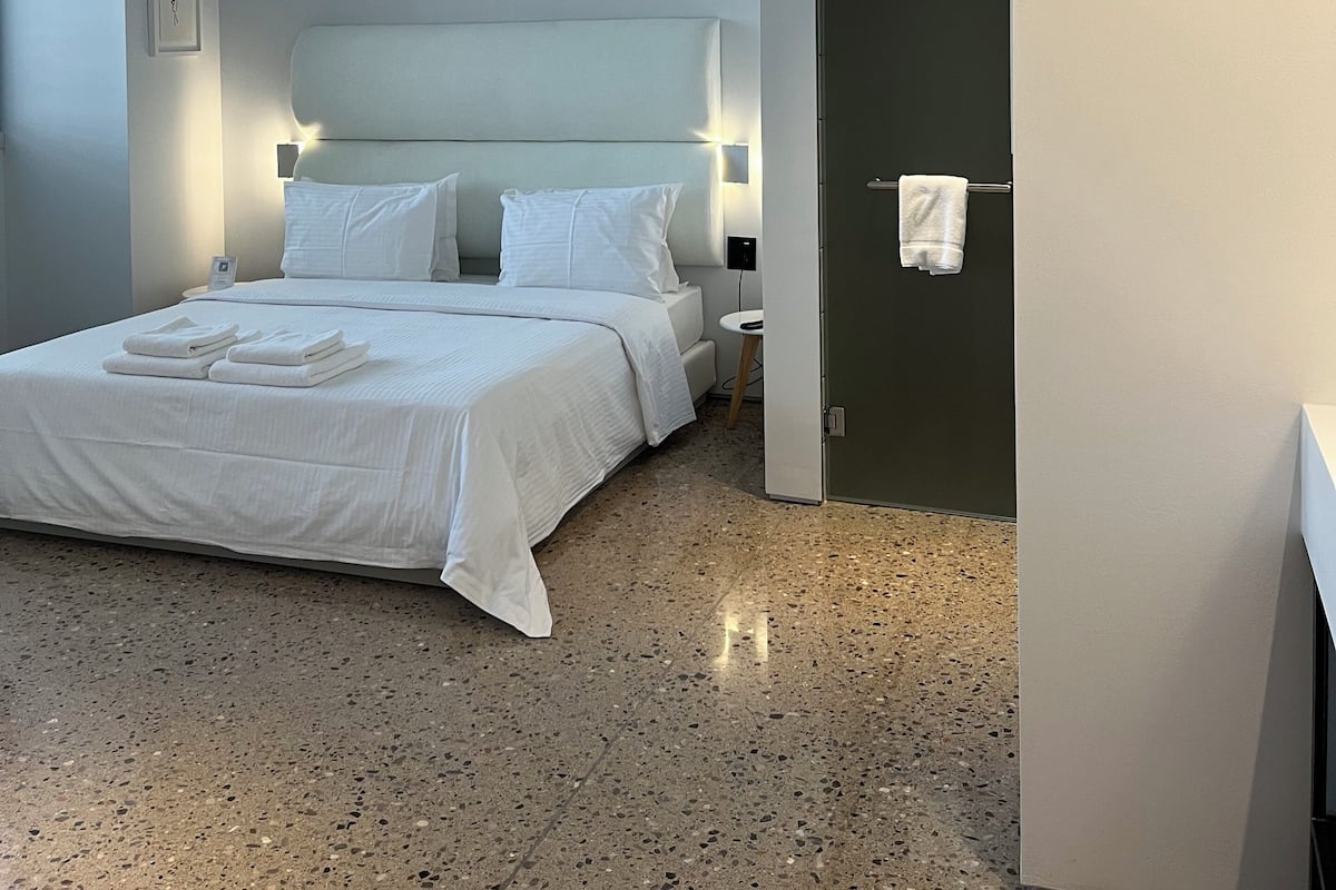 A modern hotel room with a bed, bedside tables, a nightstand, a towel rack, and a door.