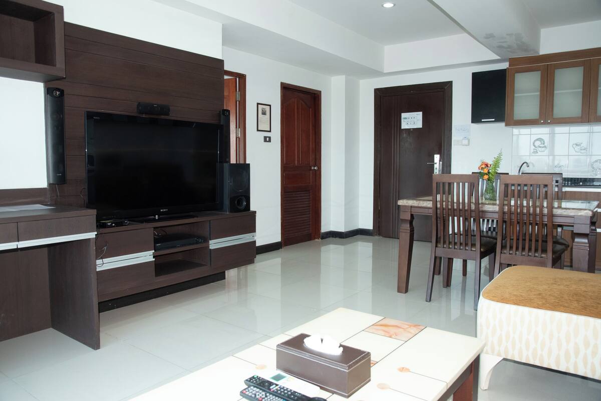 A living room with a flat-screen TV, wooden entertainment unit, dining table with chairs, and a kitchen area with cabinets.