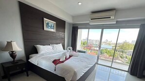 A hotel room with a large bed, a nightstand, a lamp, a picture on the wall, and a view of the city through large windows. - Valhalla Pattaya Hotel (Pattaya)