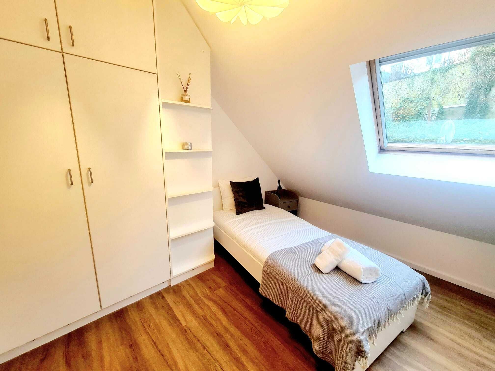 A bedroom with a bed, a wardrobe, a shelf unit, a window, and a wooden floor.