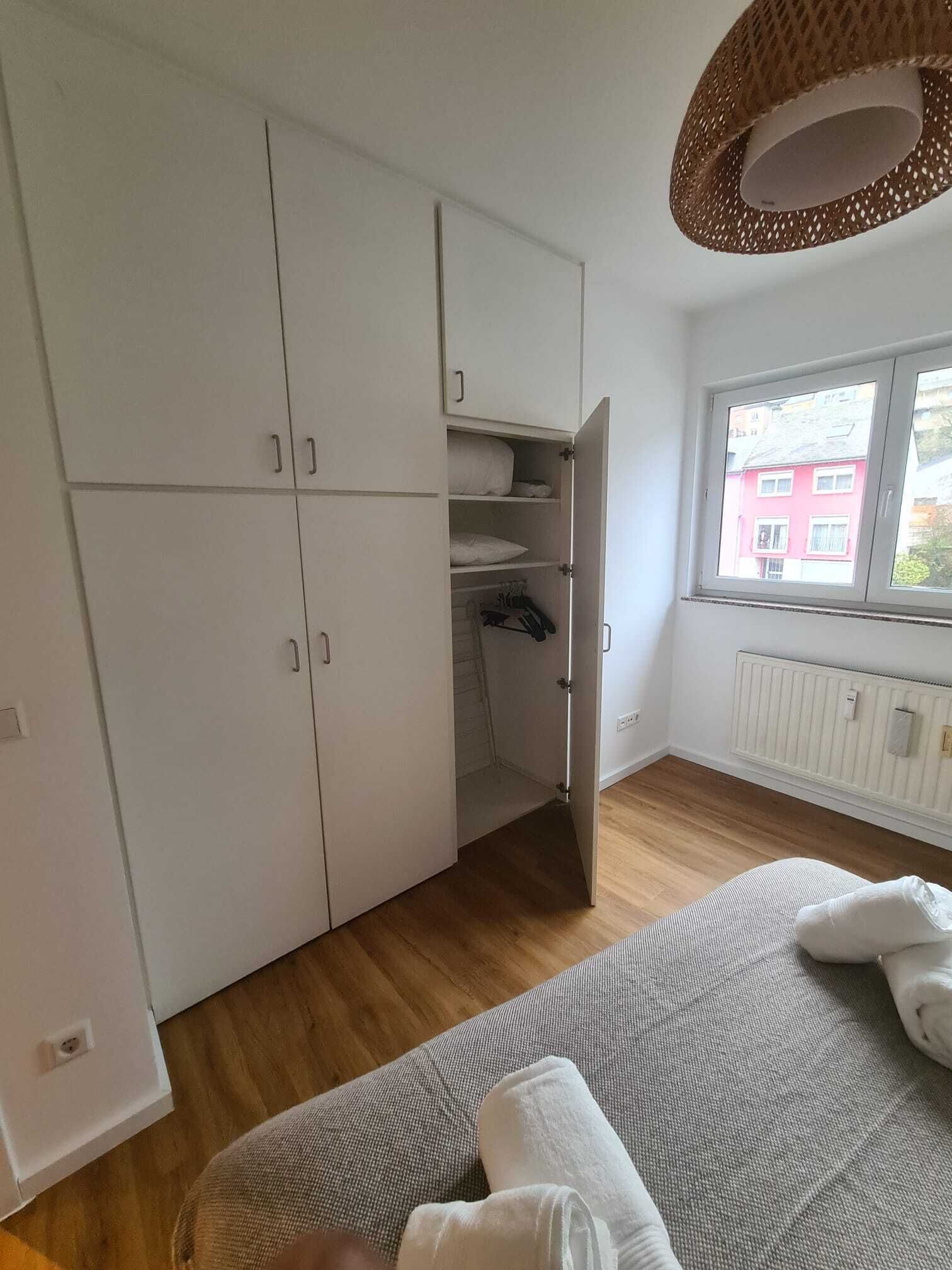 Une pièce avec une grande armoire, une fenêtre donnant sur des immeubles et un canapé gris sur lequel est posée une serviette blanche enroulée.