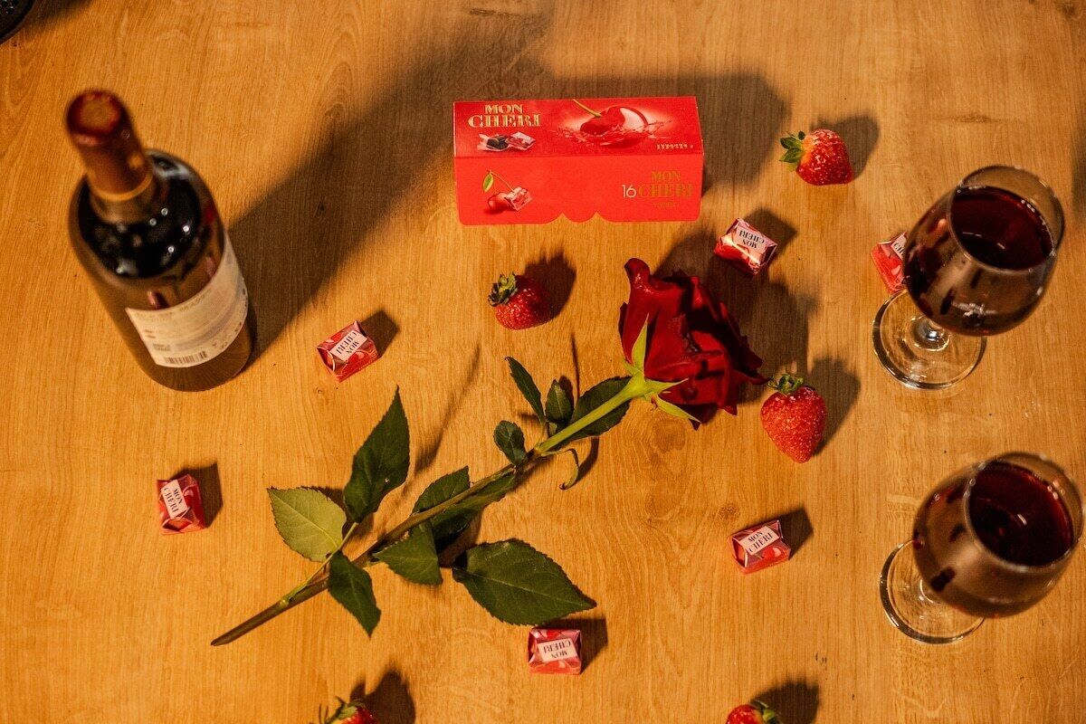 Una mesa de madera con una botella de vino, una copa de vino tinto, una caja de bombones Mon Cheri, fresas y una rosa.
