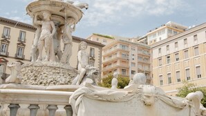 Une sculpture de fontaine classique, représentant plusieurs personnages, entourée d’une balustrade et se dressant sur fond d’immeubles d’habitation.